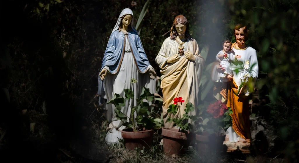 Reprise de la photo de couverture : photographie représentant trois statues religieuses installées en extérieur, disposées de gauche à droite : la Vierge Marie vêtu d&rsquo;un manteau bleu, le Sacré-Cœur de Jésus et Saint Joseph tenant l&rsquo;Enfant Jésus dans ses bras. Des pots de fleurs avec des géraniums sont posés au pied des statues dans un cadre verdoyant et ombragé.