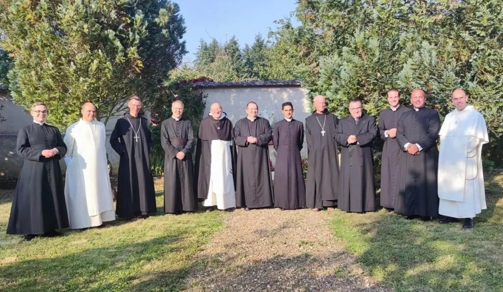Groupe d&rsquo;une douzaine d&rsquo;ecclésiastiques et de religieux représenatnt divers courants des communautés catholiques traditionalistes, posant en rangée en extérieur, dans un jardin bordé d&rsquo;arbres et d&rsquo;un mur en arrière-plan. Les membres portent divers habits religieux traditionnels, notamment des soutanes noires, des habits monastiques bénédictins noirs et dominicains blancs, ainsi que des croix pectorales pour certains évêques ou abbés. Mgr Gilles Wach figure au centre du groupe en soutane noire.