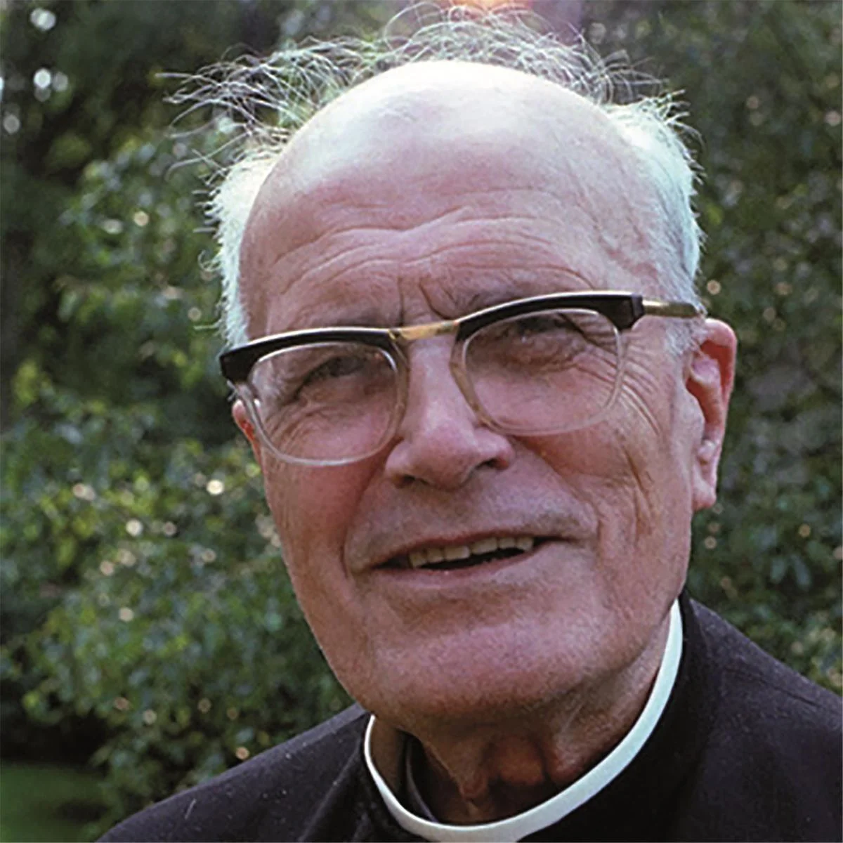 Portrait rapproché du père Ludovic-Marie Barrielle portant des lunettes et col romain, sur fond de végétation.