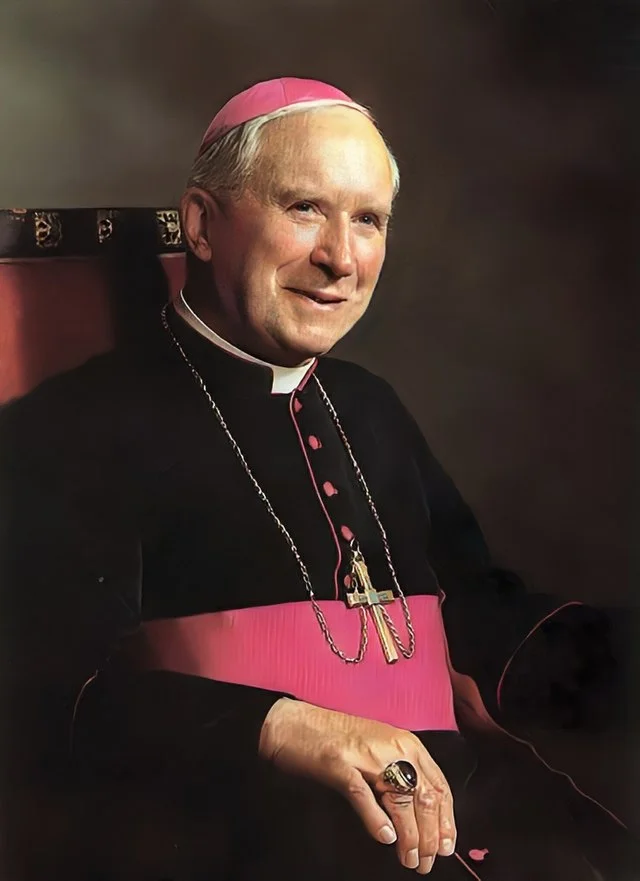 Portrait de Mgr Marcel Lefebvre en tenue d&rsquo;évêque avec calotte et ceinture violettes, portant une croix pectorale et souriant assis sur un siège.